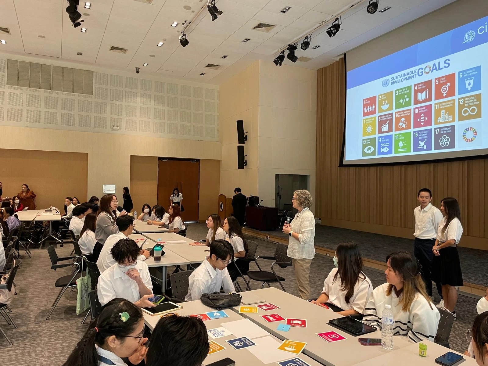 Heather Ranson teaching undergraduate students of the "Developing a Sustainability Mindset" course at Mahidol University in Bangkok, Thailand. The instructor stands in front of a large number of students sat ar tables in a large lecture hall, with a projector behind showing the UN Sustainable Development Goals.