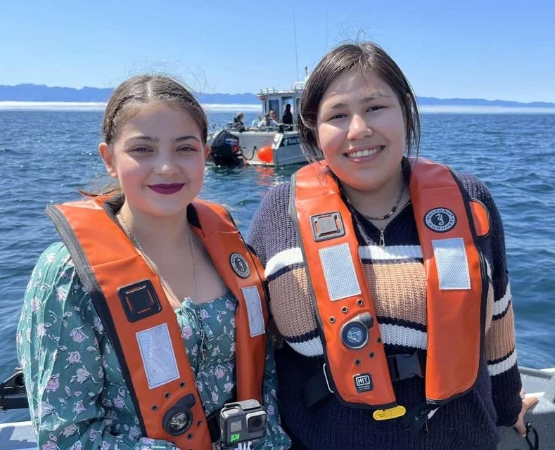 Eriel and Carmen, young Inuit leaders from Tuktoyaktuk, Northwest Territories, pose together in a boat with ocean and another boat behind, while learning about coastal stewardship at with T'Sou-ke First Nation on Vancouver Island.