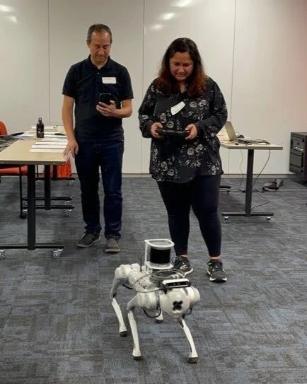 Two people standing while controlling a four-legged robot during an event held by Hikma Collective.
