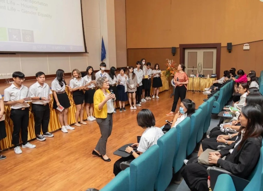 Heather Ranson teaching undergraduate students of the "Developing a Sustainability Mindset" course in a large lecture hall at Mahidol University in Bangkok, Thailand. A group of students stand against a wall below a projector screen, while the instructor speaks to the rest of the audience sat in lecture theatres seats.