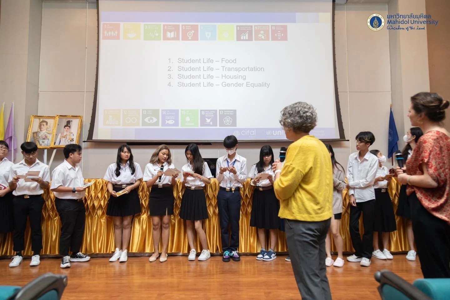 Heather Ranson teaching undergraduate students of the "Developing a Sustainability Mindset" course in a large lecture hall at Mahidol University in Bangkok, Thailand. Students stand against a wall below a projector screen, while the instructor speaks to them through a microphone.