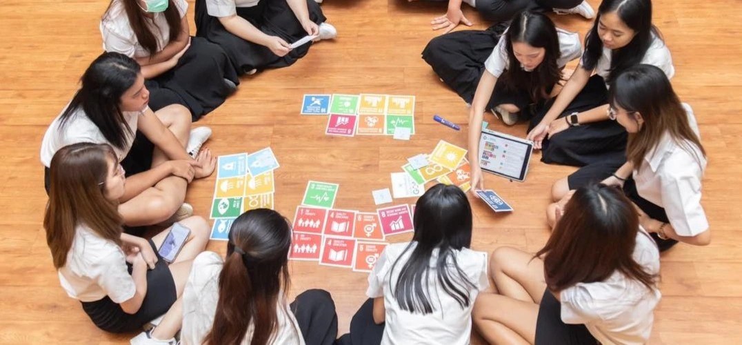 Students sat on the ground in a circle looking at cards showing the symbols representing the UN Sustainable Development Goals spread out on the floor, 